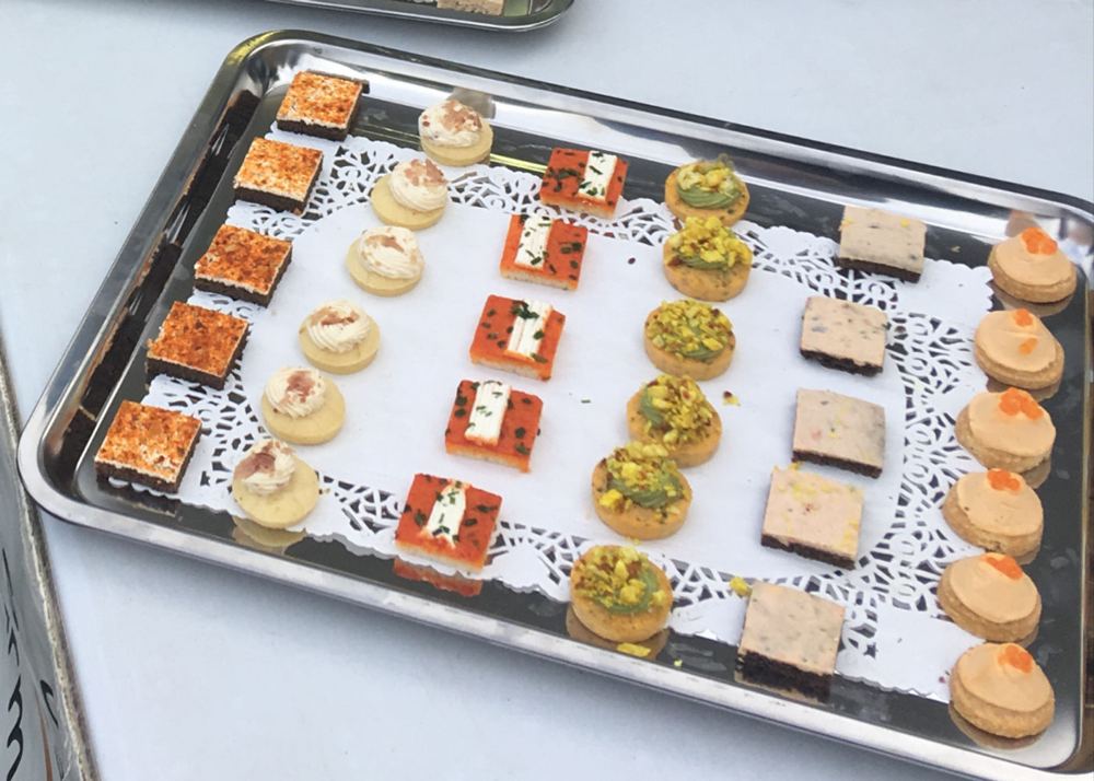 Plateau Le Grand Soir : canapés ronds et carrés en rangées, garnis de crème, d’éclats verts ou de perles orange, sur papier dentelle